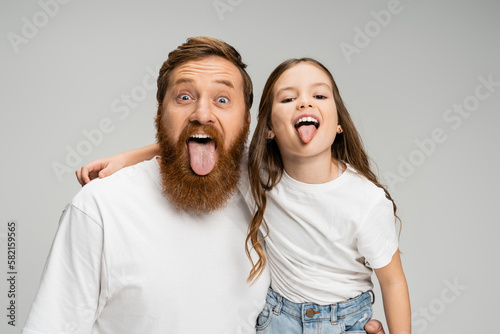Preteen kid and bearded dad sticking out tongues isolated on grey.