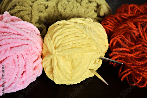 Demonstration of colored knitting threads and knitting needles sticking out of a ball of yellow threads on a dark background. Close-up
