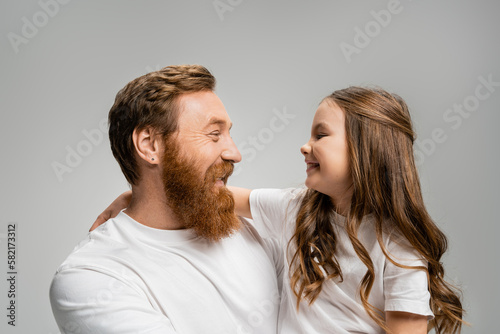 Side view of smiling girl in white t-shirt hugging bearded dad isolated on grey.