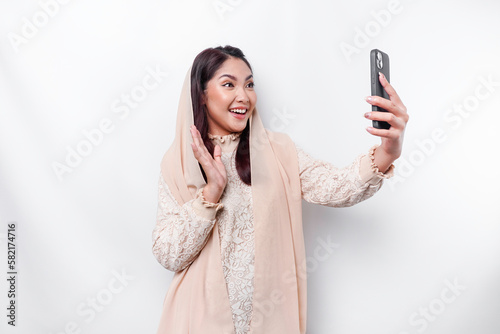 A portrait of a happy Asian Muslim woman wearing a headscarf, holding her phone, isolated by white background