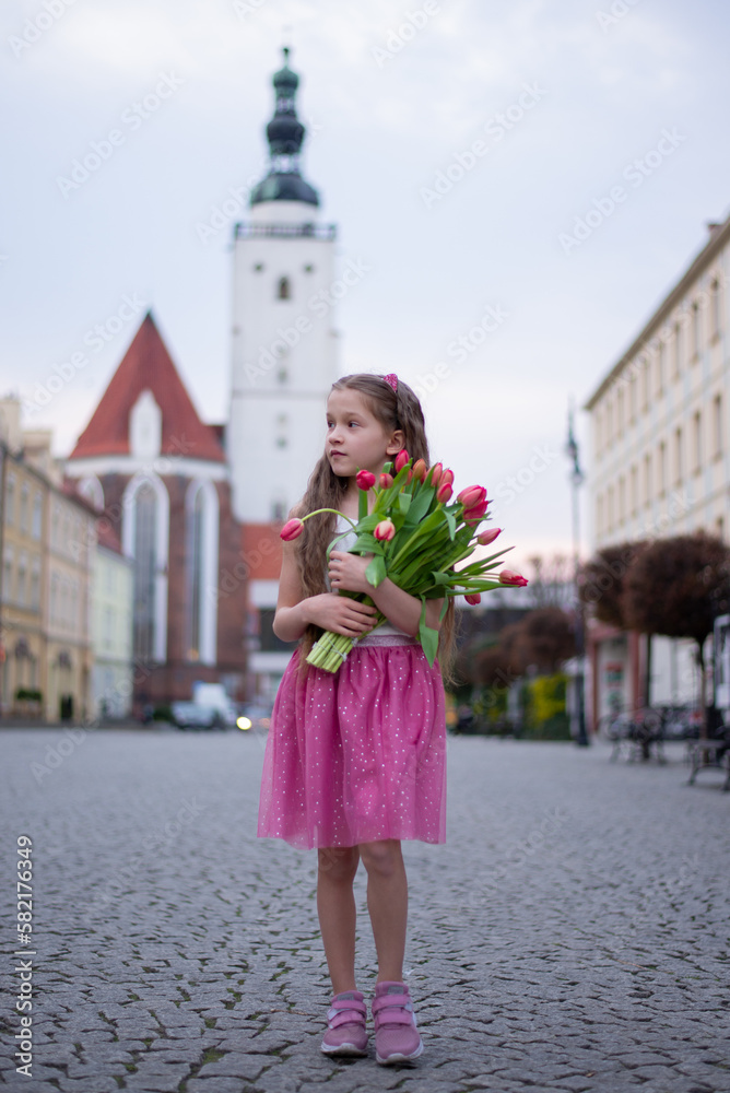 Adorable 9 years old girl with long curly hair walking through central square of the town Olesnica, Poland. Beautiful girl walking with bouquet of tulip flowers through spring European city. Adorable 9 years old girl with long curly hair walking through central square of the town Olesnica, Poland. Beautiful girl walking with bouquet of tulip flowers through spring European city.