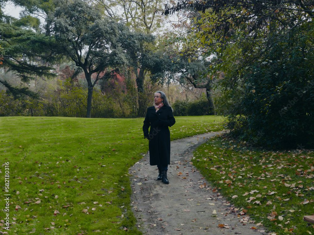 Mature woman walking along a park path. Woman in nature with green grass Mature woman walking along a park path. Woman in nature with green grass