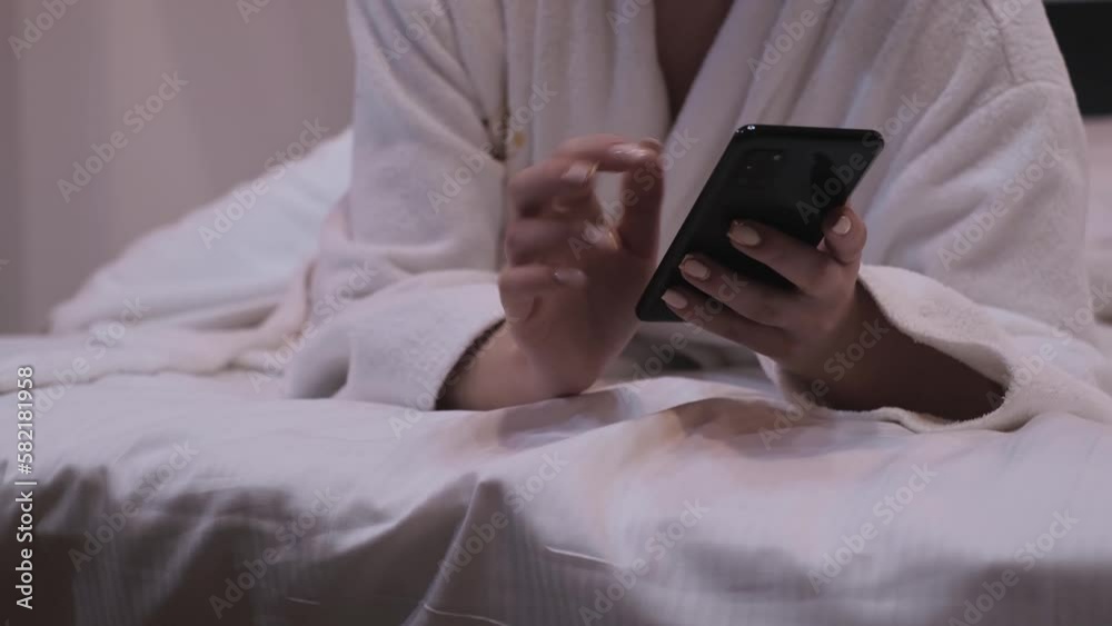 The girl works on a smartphone. A woman in a bedroom in a white bathrobe is lying in bed and typing with both hands on a laptop keyboard. Close-up The girl works on a smartphone. A woman in a bedroom in a white bathrobe is lying in bed and typing with both hands on a laptop keyboard. Close-up