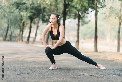 A beautiful sportswoman in sportswear is stretching her body with a smile on her face, while doing flexibility exercises to warm up before a running workout in Autumn city park background.