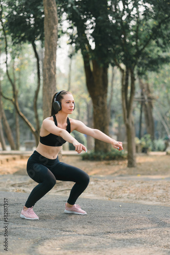 In an Autumn city park background, a stunning sportswoman in sportswear is smiling while stretching her body and performing flexibility exercises as a warm-up before a running workout.