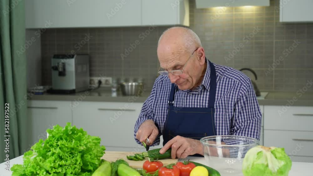 Senior man in blue apron cuts cucumber into thin slices with knife. Elderly man wants to cook salad for family. Old man sits at kitchen table Senior man in blue apron cuts cucumber into thin slices with knife. Elderly man wants to cook salad for family. Old man sits at kitchen table