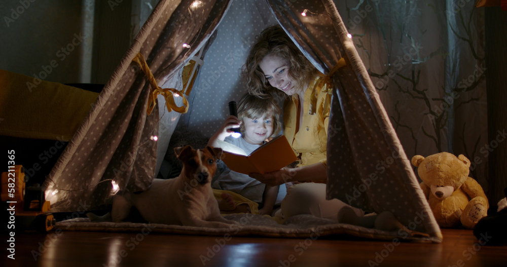 Happy caucasian mom and her cute baby boy spending time together at home, reading a book, using a flashlight in a cozy tent - happy family 
