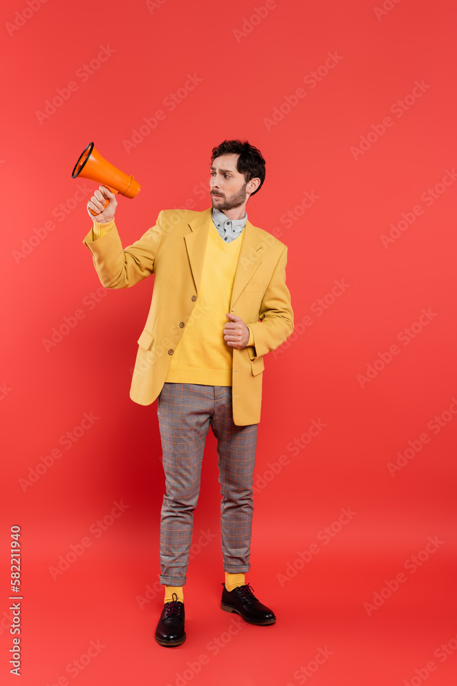 Stylish brunette man in jacket looking at loudspeaker on red background. Stylish brunette man in jacket looking at loudspeaker on red background.