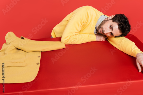 tired guy in yellow long sleeve jumper lying on red desk near blazer isolated on coral background.