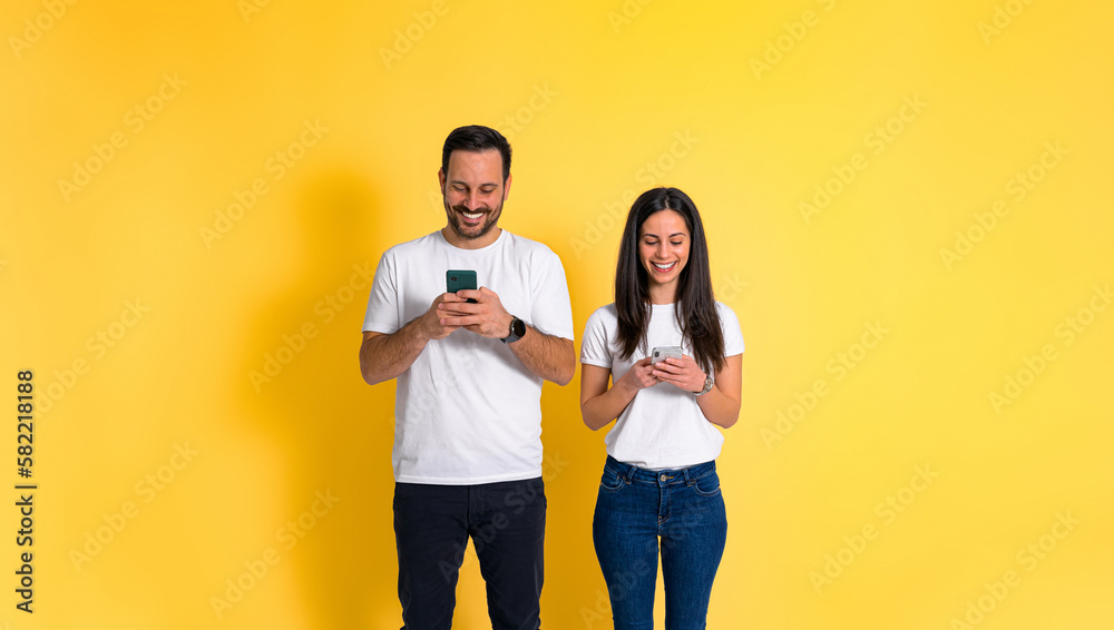 Portrait of smiling young adult couple dressed in white t-shirts text messaging and using smart phones while standing isolated on yellow background