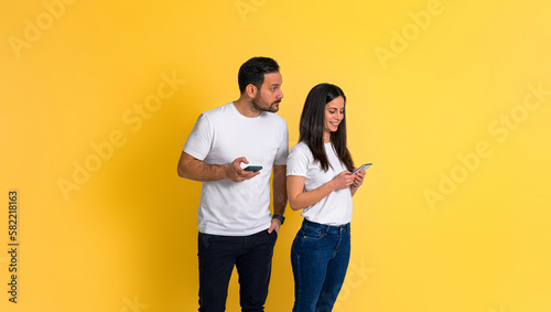 Suspicious and possessive boyfriend with mobile phone spying on smiling girlfriend chatting on social media using smartphone while standing isolated on yellow background photo