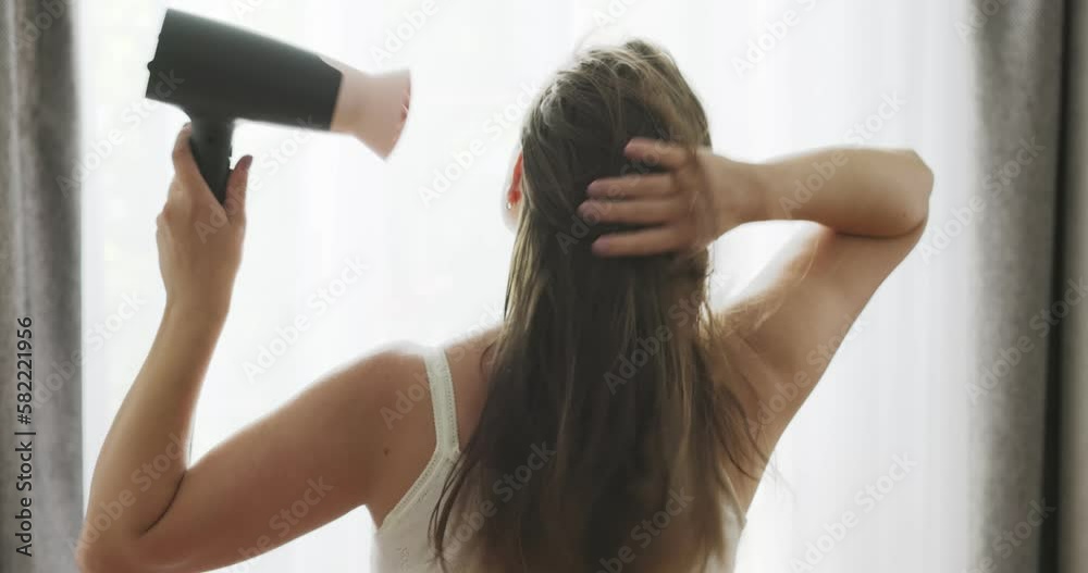 Beautiful young woman using hair dryer near the window at home