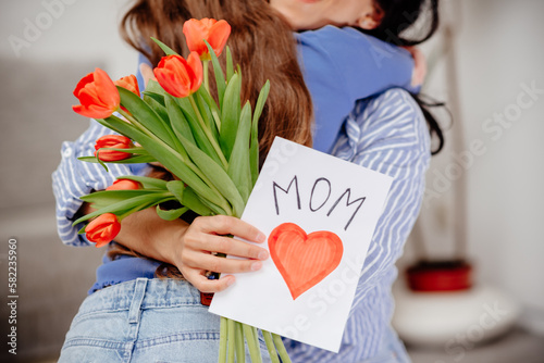 Grateful mom hugging daughter girl, holding flowers bouquet, receiving hand drawn greeting card with loving heart from girl, smiling at camera. Mothers day, 8 march, concept. Head shot portrait