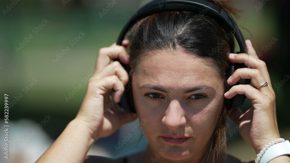 Woman in 30s putting noise cancelling headphones outdoors. Female person wearing over the ear headphone at park Woman in 30s putting noise cancelling headphones outdoors. Female person wearing over the ear headphone at park