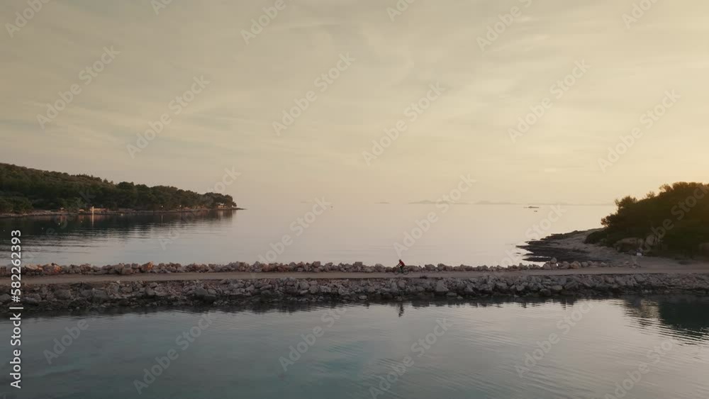 Aerial view of man riding his bicycle along the coastline at golden hour. Beautiful calm sea and orange sunset. Active lifestyle. Aerial view of man riding his bicycle along the coastline at golden hour. Beautiful calm sea and orange sunset. Active lifestyle.