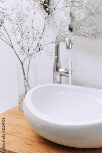 Beautiful white flowers in vase near modern sink in bathroom, water pouring on background