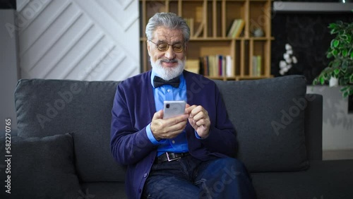 An adult man in a shirt with a bow tie holding a phone in his hand is sitting on a sofa, casually relaxed. The elderly man smiles, scrolls the phone, reads the news at home