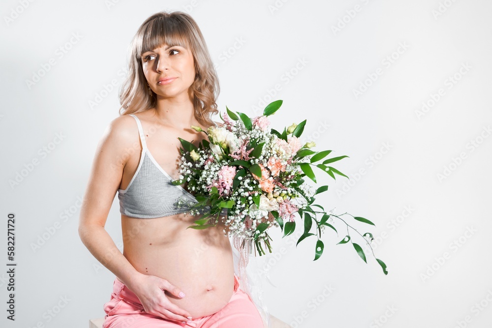 Pregnant woman with bouquet of flowers. Girl in gray top, pink pants holds hands on naked belly. Photoshoot of happy pregnancy, maternity, preparation. Baby expectation. Beautiful tender young mother Pregnant woman with bouquet of flowers. Girl in gray top, pink pants holds hands on naked belly. Photoshoot of happy pregnancy, maternity, preparation. Baby expectation. Beautiful tender young mother