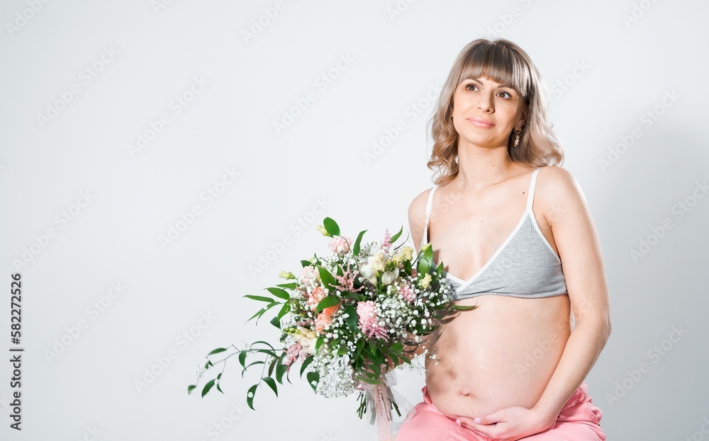 Pregnant woman with bouquet of flowers. Girl in gray top, pink pants holds  hands on naked belly. Photoshoot of happy pregnancy, maternity,  preparation. Baby expectation. Beautiful tender young mother Stock Photo