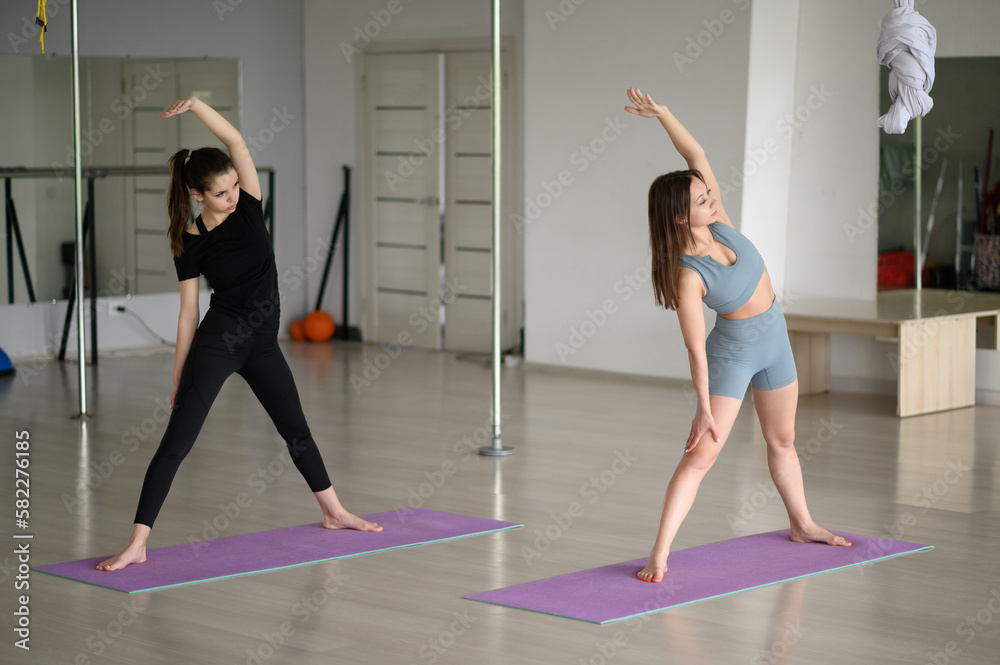 Two young Caucasian women in sportswear are doing stretching in the gym. Body tilt.The concept of a healthy lifestyle, Pilates, sports. Athletes on the way to a stronger body