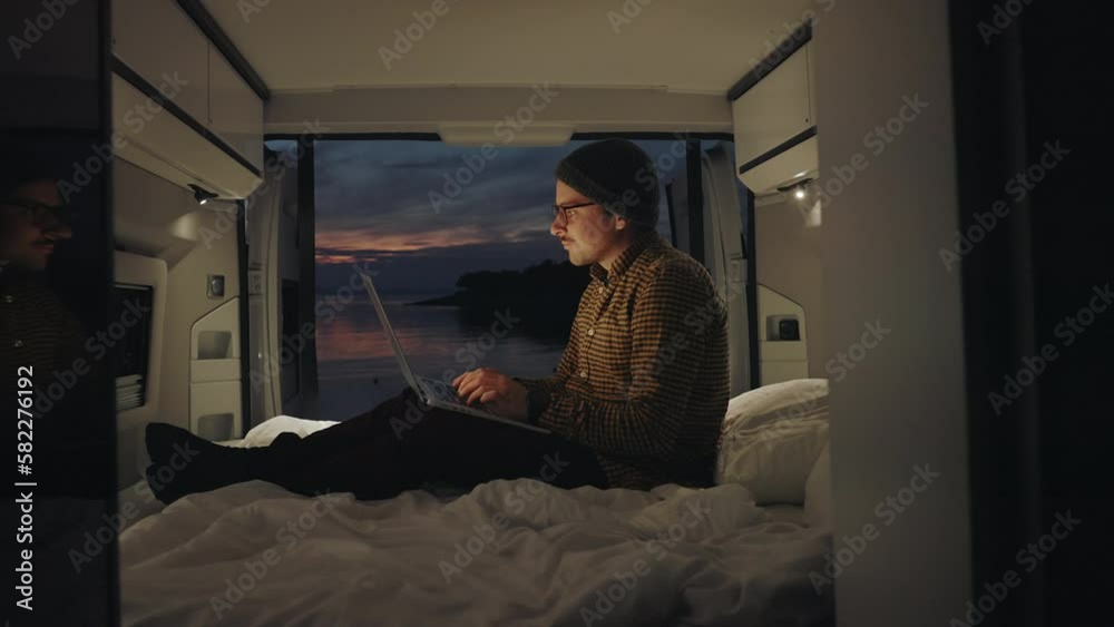 Urban digital nomad of millennial generation working on a laptop in the camper van by the seaside at sunset. Creative working inspired by the nature. Remote office job. 