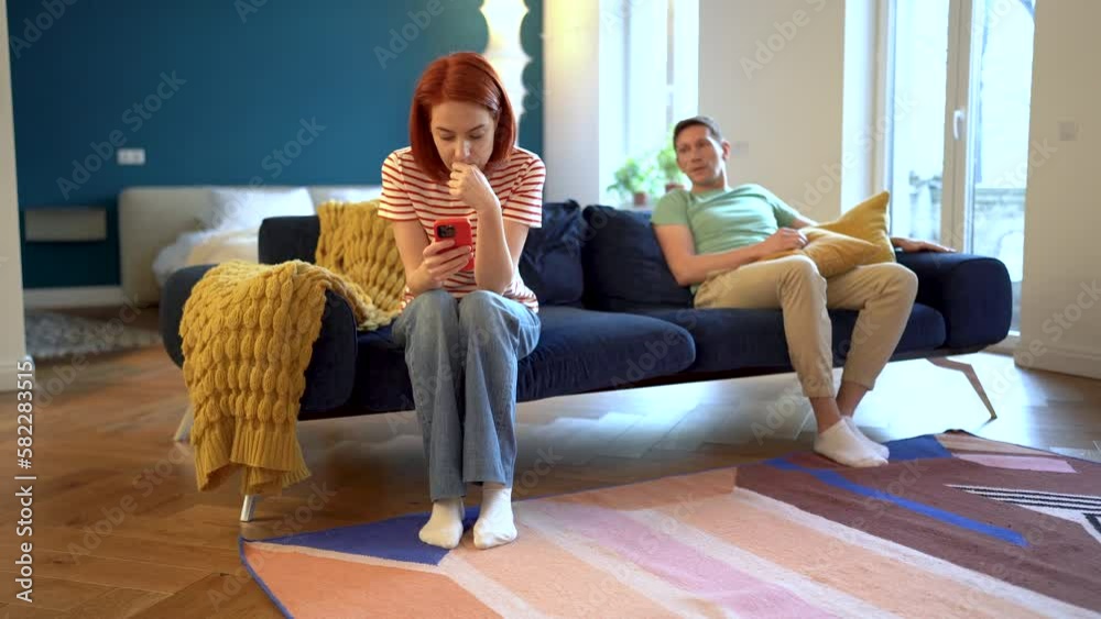 Offended woman after quarrel with husband sits stooped on couch, ignoring looks at screen smartphone smiling man wants to make peace flirting throwing sofa cushion at upset wife attracting attention Offended woman after quarrel with husband sits stooped on couch, ignoring looks at screen smartphone smiling man wants to make peace flirting throwing sofa cushion at upset wife attracting attention