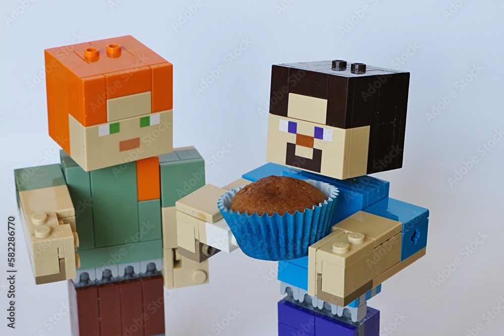 Fototapeta premium LEGO Minecraft larger figures of Steve and Alex, Steve is holding small, bronze baked muffin cake in a blue paper cup. Light grey background.