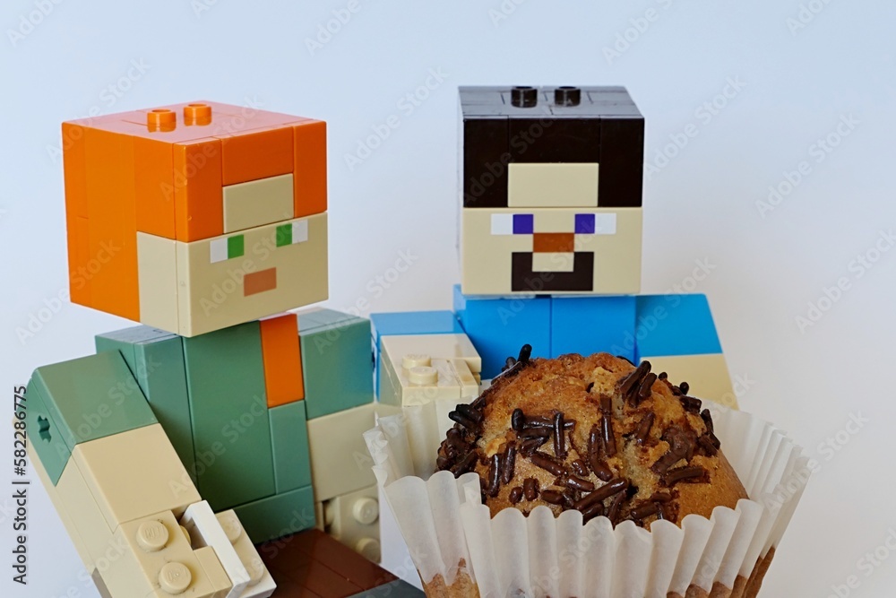 Fototapeta premium LEGO Minecraft large figures of Steve and Alex, Steve is holding huge muffin cake with chocolate chips on top in a white paper cup. Light grey background.