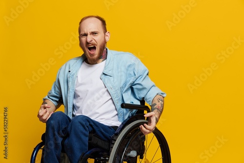 man in a wheelchair screams angrily, shows fist looks at the camera is not pleased, with tattoos on his hands sits on yellow background of the studio, the concept of health is person with disabilities