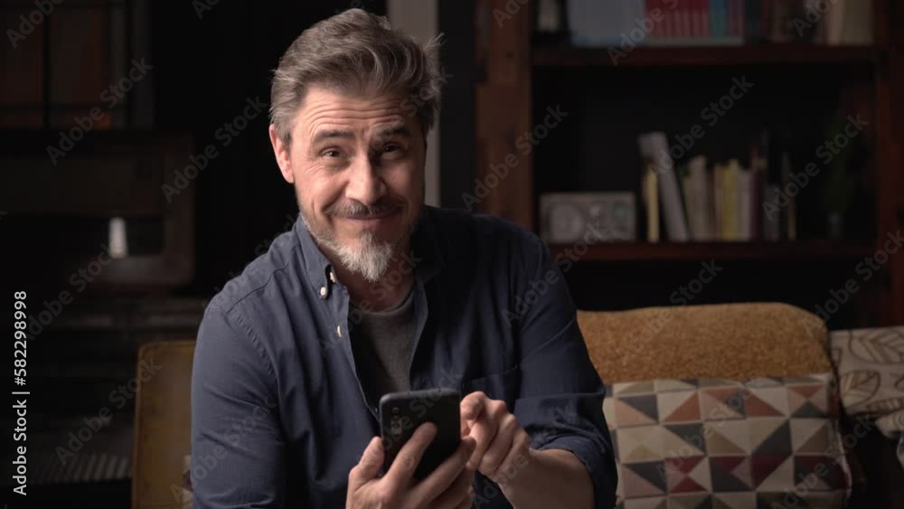 Man sitting on couch at home using phone. Businessman working in home office. Social media, newsfeed, chat. Mature age, middle age, mid adult man, bearded, authentic look. Man sitting on couch at home using phone. Businessman working in home office. Social media, newsfeed, chat. Mature age, middle age, mid adult man, bearded, authentic look.