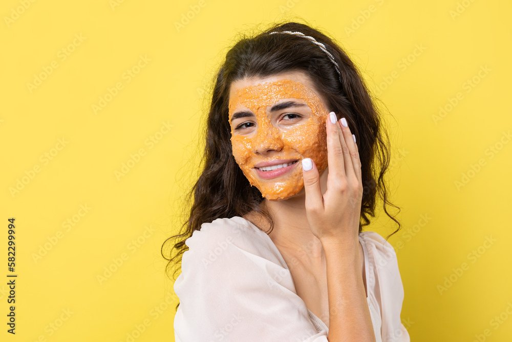 A young brunette girl with a cleansing scrub mask on her face, looks at the camera and smiles, against a yellow background. A young brunette girl with a cleansing scrub mask on her face, looks at the camera and smiles, against a yellow background.