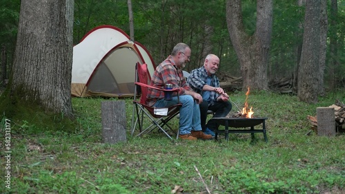 Wide angle view of two gay men building a campfire in a forest talking and laughing with tent in background. © Robert Peak