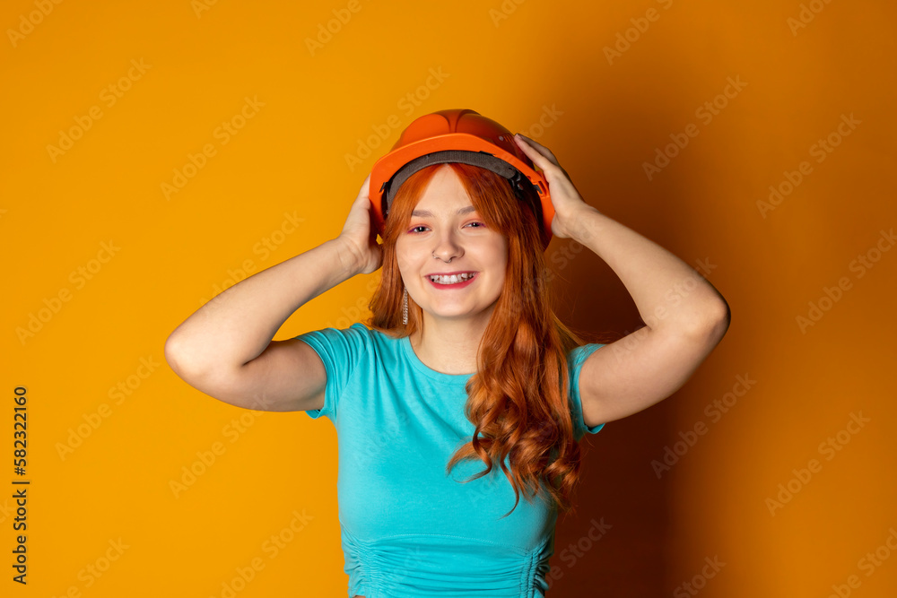 A red-haired girl wearing a construction helmet is showing a thumbs-up on an isolated orange background. Emphasizing safety regulations. A red-haired girl wearing a construction helmet is showing a thumbs-up on an isolated orange background. Emphasizing safety regulations.