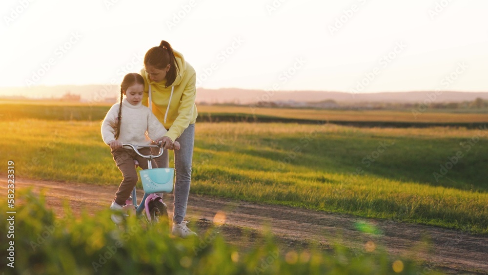 mother teaches child ride two-wheeled bicycle sunset. fun weekend by bike. child's dream kid. child drive bicycle. mom rides girl child daughter bikerays sunset. happy family concept. teamwork