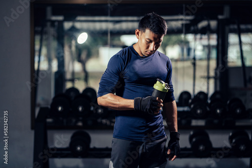 Fitness man in sportswear holding a bottle water or protein shake for drink in fitness gym. Health care and workout. Asian man replenishing water balance after workout. Concept of health and wellness.