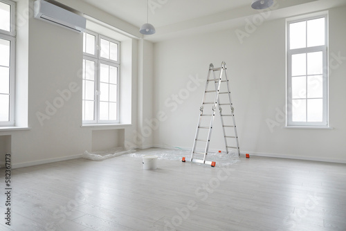 Modern apartment after renovations. New empty office interior or living room at home with white freshly painted walls, step ladder, paint bucket, plastic on the floor, big windows, and air conditioner