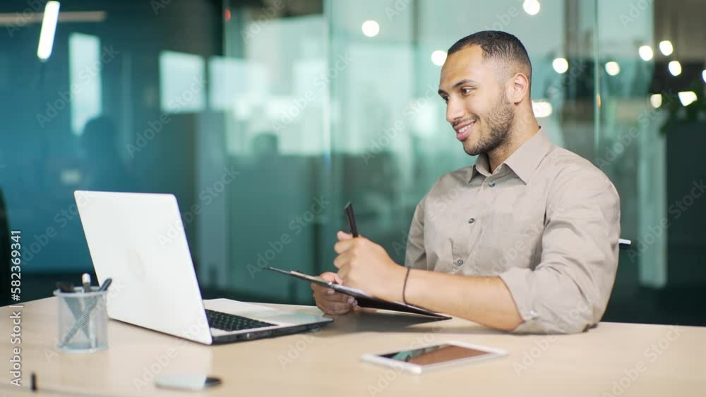 Smiling young businessman talking on a video call and writing in a notebook while sitting at a workplace at a desk in a modern office. Happy confident mixed race entrepreneur having online chat Smiling young businessman talking on a video call and writing in a notebook while sitting at a workplace at a desk in a modern office. Happy confident mixed race entrepreneur having online chat