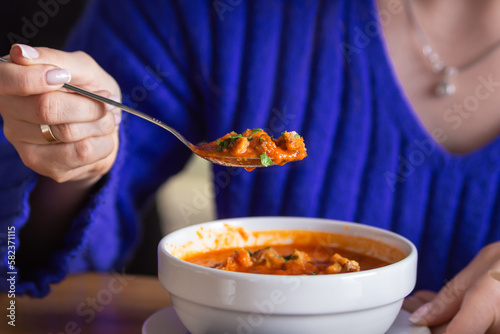 Woman eating tasty tomato soup gazpacho with croutons, vegetables and herbs in cafe. Hand holds a full spoon over a bowl