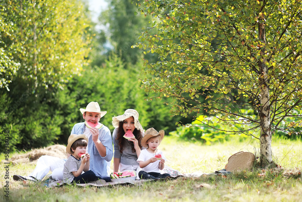 Happy family with children having picnic in park, parents with kids sitting on garden grass and eating watermelon outdoors Happy family with children having picnic in park, parents with kids sitting on garden grass and eating watermelon outdoors