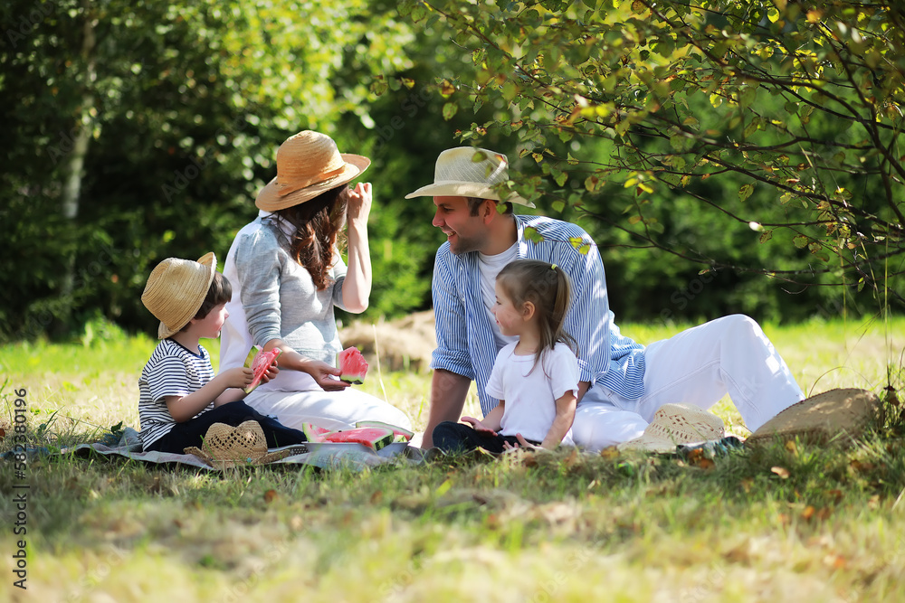 Happy family with children having picnic in park, parents with kids sitting on garden grass and eating watermelon outdoors Happy family with children having picnic in park, parents with kids sitting on garden grass and eating watermelon outdoors