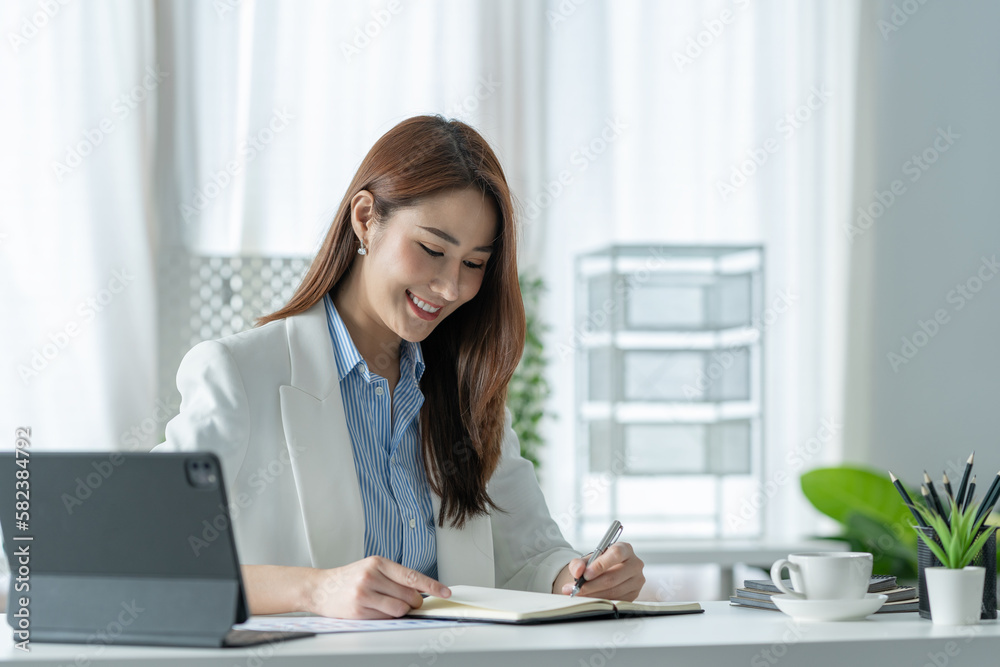 Confident Asian businesswoman sitting and taking notes in financial book Income tax with laptop computer or tablet in a happy office. Confident Asian businesswoman sitting and taking notes in financial book Income tax with laptop computer or tablet in a happy office.