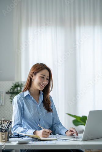 Confident Asian businesswoman sitting and taking notes in financial book Income tax with laptop computer or tablet in a happy office.