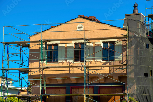 Adobe style house design with shutters and double gable roof under construction with metal scafolding in sun blue sky