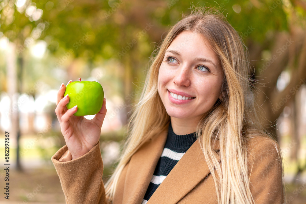 Young pretty blonde woman with an apple at outdoors looking up while smiling Young pretty blonde woman with an apple at outdoors looking up while smiling