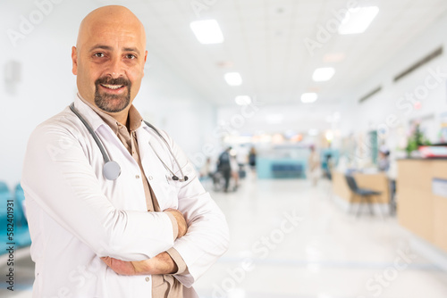 Trusted male doctor, portrait of proud middle aged trusted male doctor. Stands arms crossed in hospital. Blurred abstract medical center background with copy space. Looking camera, wearing white coat.