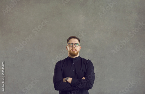 Smart businessman with folded hands on chest thinking looking up standing over studio grey background. Worry pensive man in eyeglasses staring upwards face portrait shot. People emotion