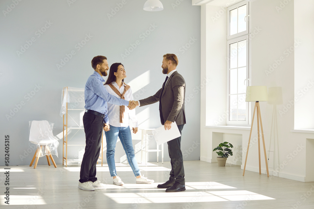 Young married couple make deal, buy new house and exchange handshake with relator. Happy man and woman standing in modern light spacious light living room and shaking hands with real estate agent - obrazy, fototapety, plakaty 