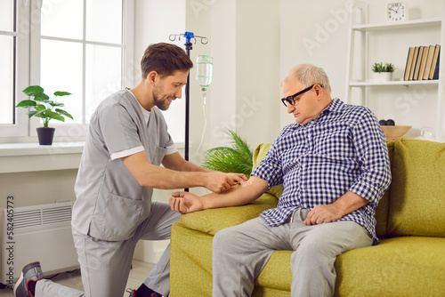 Doctor gives intravenous infusion to senior patient. Young nurse in scrubs uniform inserts IV line needle in vein of old man sitting on sofa at home. Medicine, vitamin therapy, medication concept photo