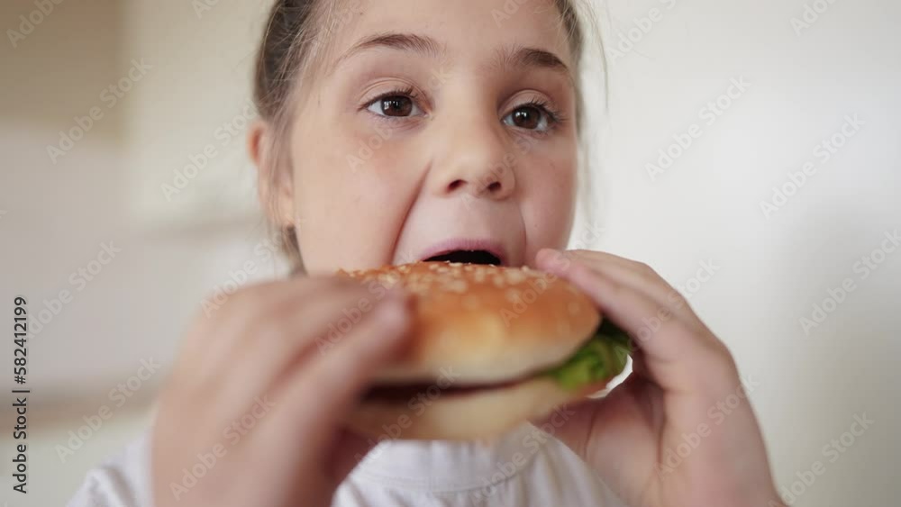 little girl eating a hamburger. unhealthy fast food proper nutrition concept. child greedily with pleasure bites a big burger in meal the kitchen at home. kid eats fast food close-up little girl eating a hamburger. unhealthy fast food proper nutrition concept. child greedily with pleasure bites a big burger in meal the kitchen at home. kid eats fast food close-up