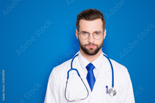 Portrait of trendy modern doc with stubble and modern hairstyle in white lab coat, tie and stethoscope on his neck, looking at camera, isolated on grey background